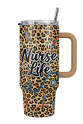 A 32oz insulated travel tumbler with a handle, featuring a leopard print design with 'Nurse Life' text, stethoscope, syringes, and EKG line graphics. It has a clear lid with a black-and-white striped straw.